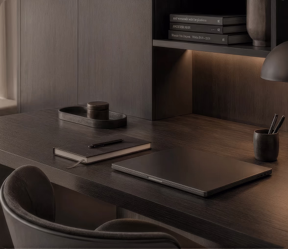 Dark wood desk with a closed laptop, a leather notebook and a lit shelf above